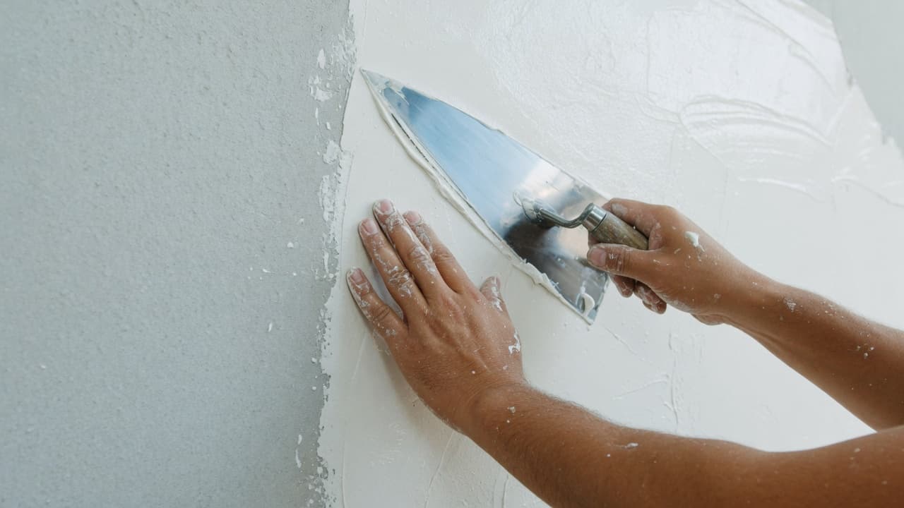 Hands smoothing fresh white plaster onto a wall with a steel trowel